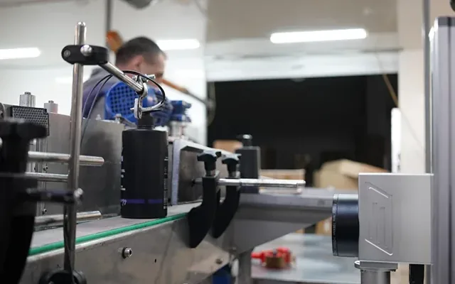 Laser marker above the green conveyor belt of a household-chemicals line, with the code inspection camera beside it