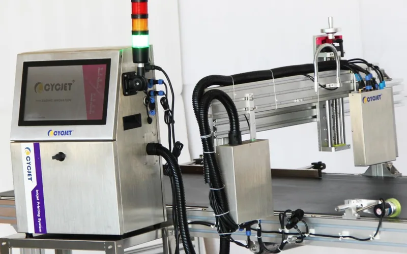 An inkjet printer beside a conveyor: control unit with a screen, ink supply hoses and the print head above the belt